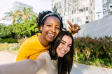 Fototapeta Panele - Two young beautiful women taking selfie portrait together in summer at city. Diverse girls enjoying free time outdoors. Female friendship concept.