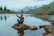 © Anna Tsukanova/Stocksy - Man sitting at scenic lake at using smartphone to take a picture