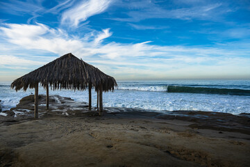 Wall Mural - 2023-12-31 A GRASS HUT STRUCTURE ON A ROCKY SHORELINE WITH WAVE COMING TO SHORE AND A INTENSE BLUE SKY WITH NICE HEITE COUDS AT THE WINDANSEA BEACH IN LA JOLLA CALIFORNIA