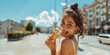 Funny teenage girl or young woman looking over her shoulder while licking melting ice cream on hot summer street, tongue visible, copy space on blurred city background
