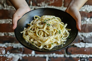 Wall Mural - Hands Holding Black Plate with Creamy Fettuccine Alfredo, Brick Wall Background