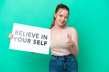 Canvas Print - Young caucasian woman isolated on green background holding a placard with text Believe In Your Self and pointing to the front