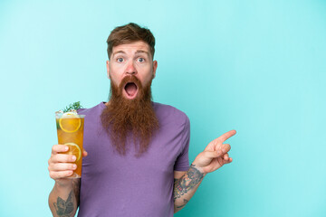 Wall Mural - Redhead man with long beard holding a cocktail isolated on blue background surprised and pointing finger to the side