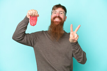 Wall Mural - Redhead man with long beard holding a piece of meat isolated on blue background smiling and showing victory sign