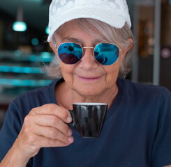 Poster - Close up portrait of attractive senior woman with blue sunglasses sitting at cafe table holding an espresso coffee cup