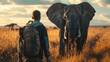 A man is walking in the desert with a backpack and an elephant is in the background. The man is looking at the elephant and seems to be in awe of its size. The scene is peaceful and serene