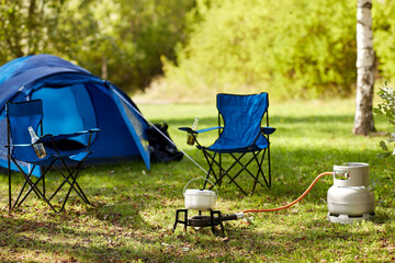 food cooking, tourism and travel concept - close up of camping pot on tourist gas burner, blue tent and two folding chairs at camp