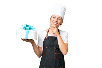 Wall Mural - Young pastry blonde woman with a big cake over isolated chroma key background looking to the side and smiling