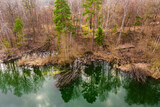 Fototapeta Na ścianę - Położone wśród lasów jezioro, którego woda ma szmaragdowy kolor. Brzegi pokrywają żółte, suche trawy, bezlistne drzewa, między którymi widać zielone korony drzew iglastych. 