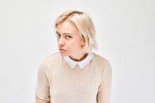 Skeptical Young Woman With Short Blonde Hair, Wearing A Beige Sweater Over A Collared Shirt, Glancing Sideways Against A White Background.