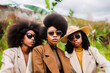 Three stylish women with afro hairstyles wearing chic sunglasses and hats in an outdoor natural setting.
