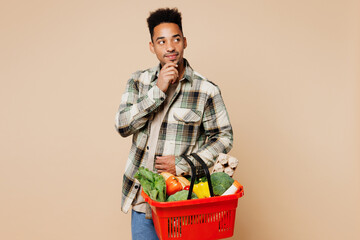 Wall Mural - Young minded pensive man he wears grey shirt hold red basket bag with food products prop up chin look aside isolated on plain pastel light beige background. Delivery service from shop or restaurant.