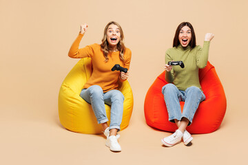 Wall Mural - Full body young friends two women wear orange green shirt casual clothes together sit in bag chair hold in hand play pc game with joystick console do winner gesture isolated on plain beige background.