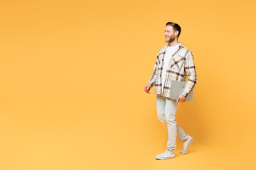 Full body side view young happy confident IT man wear brown shirt casual clothes hold closed laptop pc computer walk go isolated on plain yellow orange background studio portrait. Lifestyle concept.