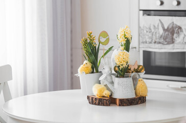 Wall Mural - Happy Easter. Spring yellow hyacinth flowers in an egg-shaped vase, chicken chicks on a white table. In the background is a white Scandinavian-style kitchen. Easter decor in the house.