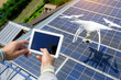 A person is holding a tablet and controlling a white drone with a camera. The drone is hovering near solar panels. Only hands in the frame