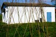 Landschaft Panorama mit grauen hohen trockenen Strauchzweigen auf grüner Weide vor weißem Betonschuppen mit braunem Dach vor blauem Himmel bei Sonne am Mittag im Frühling