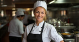Fototapeta Do pokoju - Portrait of a young female chef standing on the background of a kitchen. Smiling beautiful woman chef in the kitchen on a blurred background. Happy blonde female cook wearing a white chef's uniform.