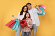 © Home-stock - Cheerful family of three people holding bright shopper bags, excited teen girl holding wrapped present box, posing isolated on yellow studio wall