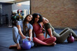© Flamingo Images - Three diverse young women sit together on the floor of a gym with kettlebells in colorful sportswear. They look confidently at the camera
