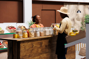 Wall Mural - Male shop owner discussing fresh fruits and vegetables at local market with a smiling black woman. Seller offers organic pantry staples and plastic-free bulk products, promoting healthy produce.