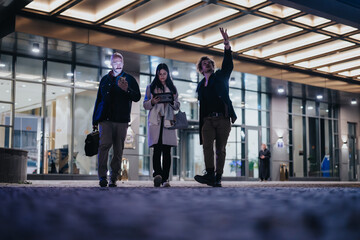 Poster - Entrepreneurs exiting an office building, engaged in an informal discussion with one person gesturing upwards, in an urban night setting.