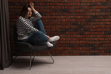 Canvas Print - Sad young woman sitting on chair near brick wall indoors, space for text