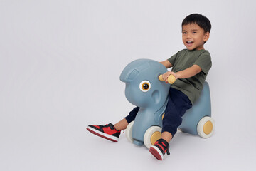 Wall Mural - Full body little small smiling happy Asian boy 2-3 years old wearing green t-shirt  sitting on toy rocking horse, playing with a toy and looking at camera isolated on white background