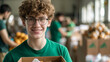 A young person in a green shirt and eyeglasses is smiling at the camera while holding a box filled with various food items, suggesting a food donation or charity event