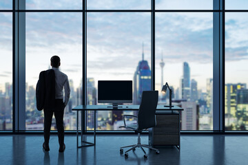 Canvas Print - Back view of young businessman standing in modern office interior with empty computer monitor on desk and panoramic windows with beautiful city view and daylight. Mock up. Workplace concept.