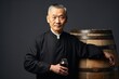 © gankevstock - An older East Asian master distiller, with a dignified presence, leans on a wooden barrel, showcasing his expertise and tradition in distillation
