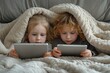 Two young children laying on a bed engrossed in technology, one holding a tablet and the other an ipad