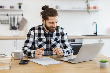 Wall Mural - Proficient Caucasian consultant receiving online conference call using technologies. Cheerful bearded adult looking at webcam of portable computer while greeting colleague in home office.