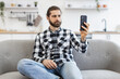 © sofiko14 - Efficient freelance worker talking to colleague while performing project for employer at kitchen. Positive Caucasian male holding smartphone while have video call, sitting at couch at home.