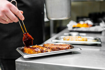 Wall Mural - Professional chef in a kitchen glazing freshly grilled ribs with a thick sauce, with other food trays in the background.