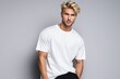 Blond, sporty European male, 25 years old, sporting a short hairstyle, docks a pose wearing an oversized white T-shirt, paired with black trousers against a neutral gray backdrop