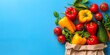 Crisp vegetables spill from a paper bag; fresh, vibrant bell-peppers and tomatoes on a blue background, with ample copyspace for healthy living concepts.