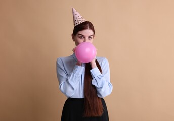 Canvas Print - Young woman in party hat blowing balloon on beige background