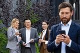 Diverse group of colleagues standing behind man with cell phone in hands and having discussion with disapproving expression. Caucasian women and asian male gossiping about irresponsible employee.