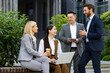 © Liubomir - Company of coworkers in formalwear resting outside of corporate building in friendly atmosphere. Two men standing by couple of women sitting on bench with coffee cup and laptop during lunch break.
