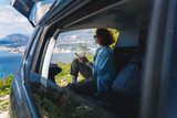 Fototapeta  - Young woman sitting in the open trunk of a car overlooking the sea with a smartphone in her hands, summer vacation and auto travel