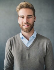 a smiling person with a beard, wearing a collared shirt under a v-neck sweater, against a neutral gray background 