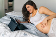 © DusanJelicic - pregnant woman with long brown hair is lying on the bed and using her tablet and phone as she does some research
