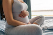 © DusanJelicic - A pregnant woman with long brown hair is sitting on the bed in front of the window with her legs crossed and her hands gently on her stomach