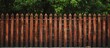 A closeup of a picket fence made of wood with trees in the background, surrounded by a grassy land lot. The fence is stained with a rich wood stain, adding charm to the home fencing