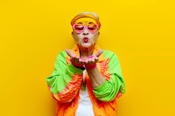 funny crazy granny in hipster clothes glasses and cap sends an air kiss on a yellow isolated background, elderly cool woman in fashionable youth clothes holds empty hands and shows flirting gesture