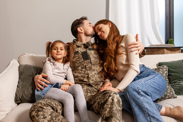 Wall Mural - Ukrainian army soldier in camouflage uniform returned home to his family, military cadet sits on the sofa with his wife and daughter and smiles, child hugs veterans father