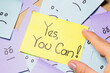 A person holds a sticky note with the message ¨Yes, you can!¨ over a pile of sticky notes with sad and angry faces. Motivational message.