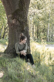 Fototapeta  - This image captures a young woman enjoying a quiet moment in the woods, seated at the base of a large tree with sunlight filtering through the leaves, creating a peaceful and reflective atmosphere