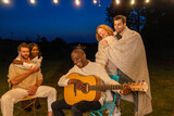 Fototapeta  - A group of friends enjoys a serene moment under the night sky, wrapped in blankets, as one plays the guitar, creating a cozy and musical outdoor experience. Cozy Nighttime Serenade at an Outdoor